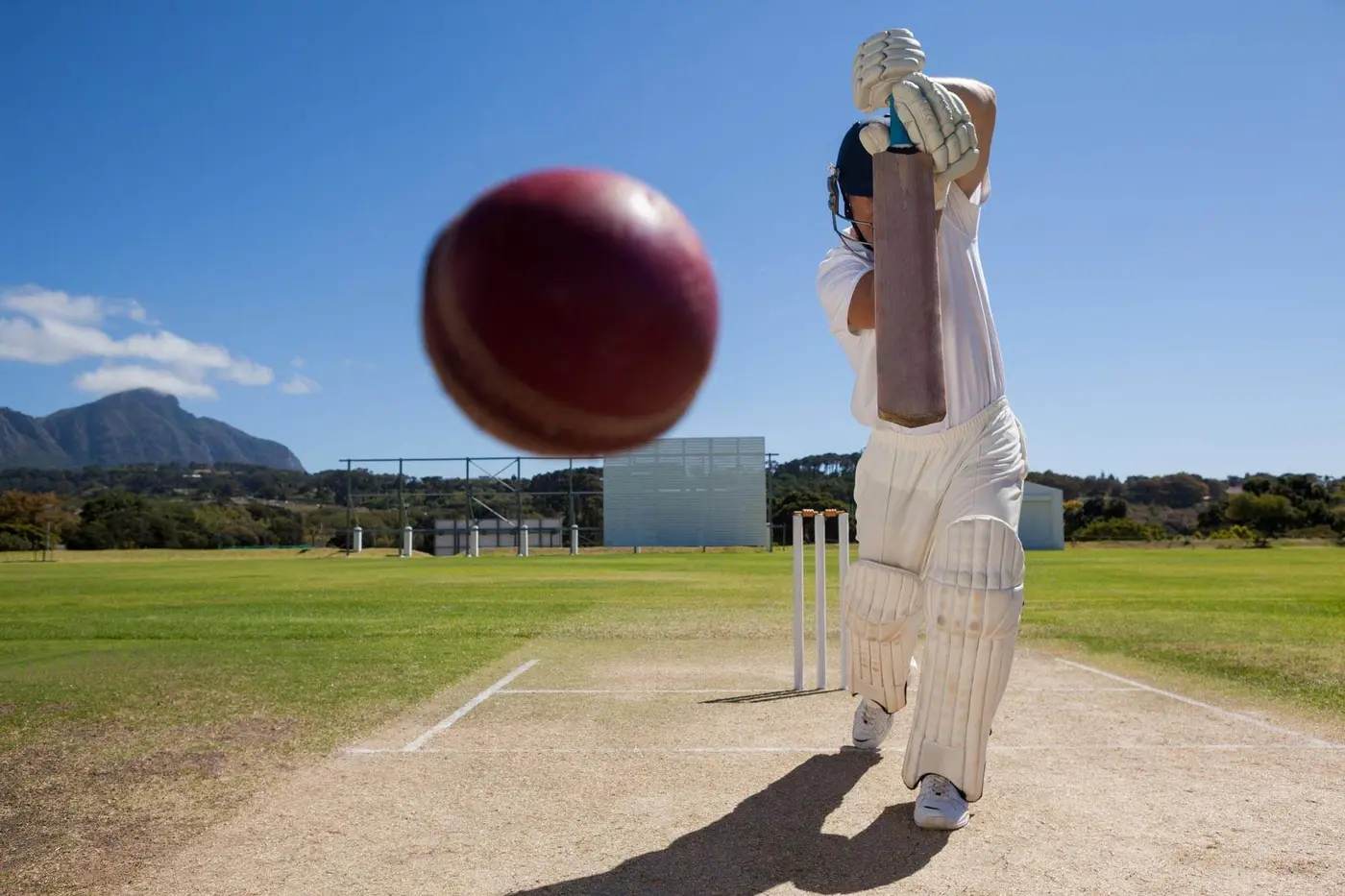 Cricket player in white pads bats on a sunny pitch using our Cricket Coaching Mat, red ball flying, mountains and blue sky behind.