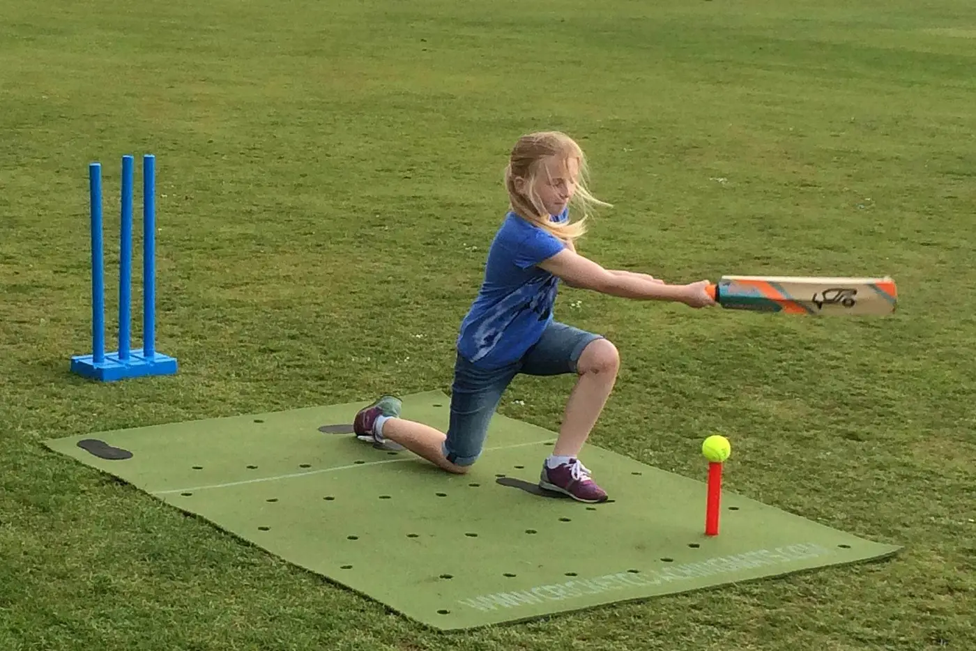 A young girl uses a Cricket Coaching Mat, swinging at a ball on a red tee, blue stumps behind her—ideal for cricket practise.